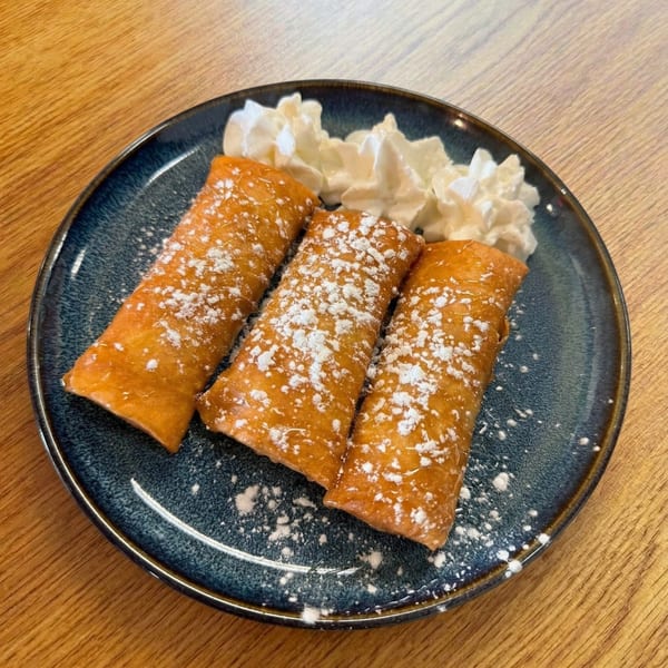 Three golden-brown dessert spring rolls dusted with powdered sugar, served with whipped cream on a blue plate.