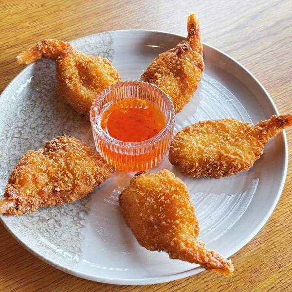Five crispy fried shrimp arranged around a small glass bowl of orange dipping sauce on a white plate.